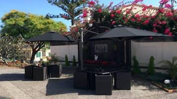 An outdoor seating area with black umbrellas and square tables, surrounded by potted plants and flowers.