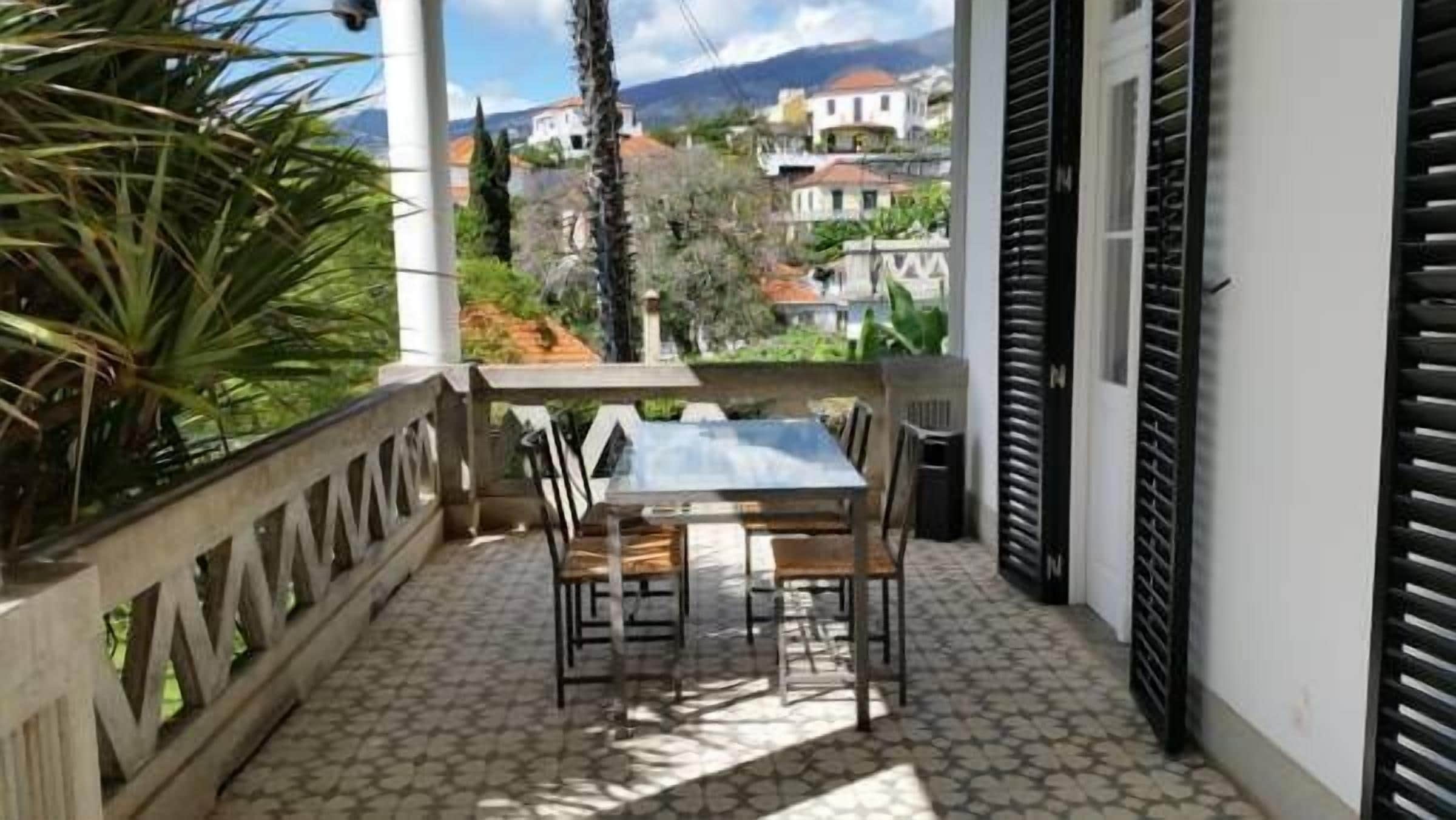 A balcony with a table and chairs, a tiled floor, and a view of houses in the background.