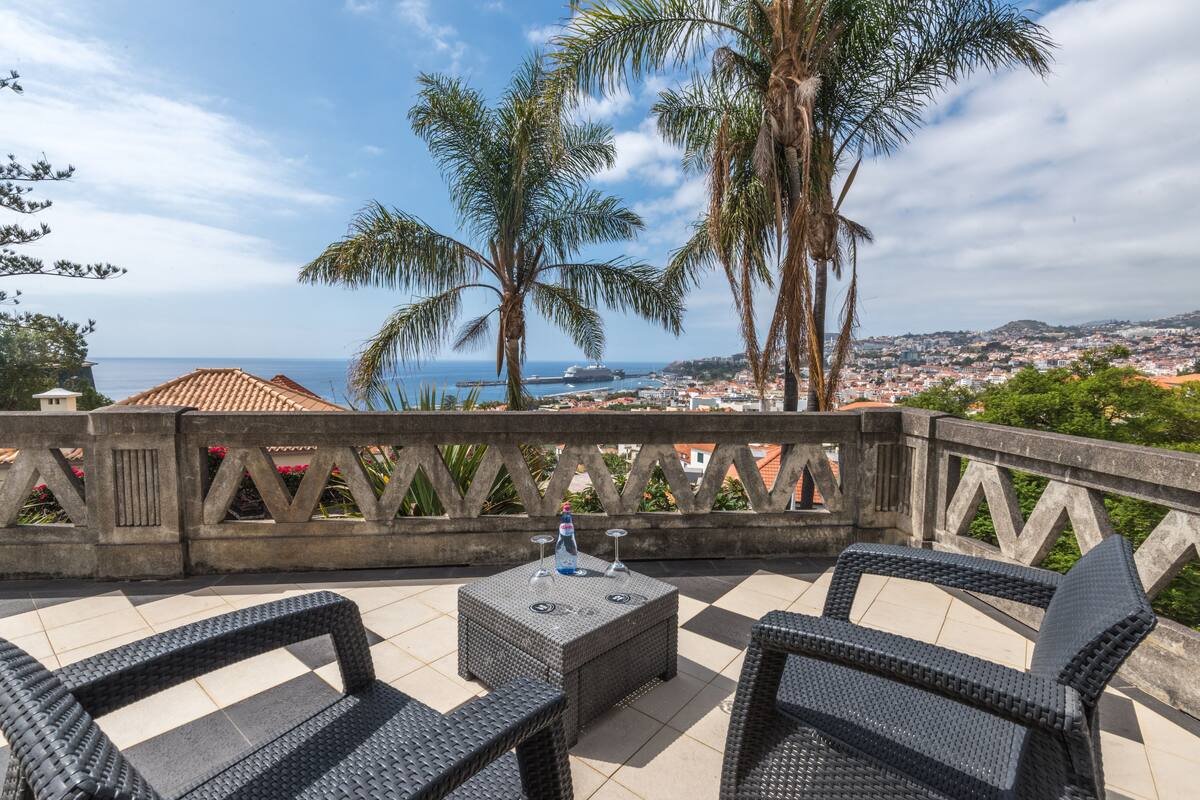 Un área de estar al aire libre con muebles de mimbre, una pequeña mesa y vista al mar y a la ciudad en el fondo.