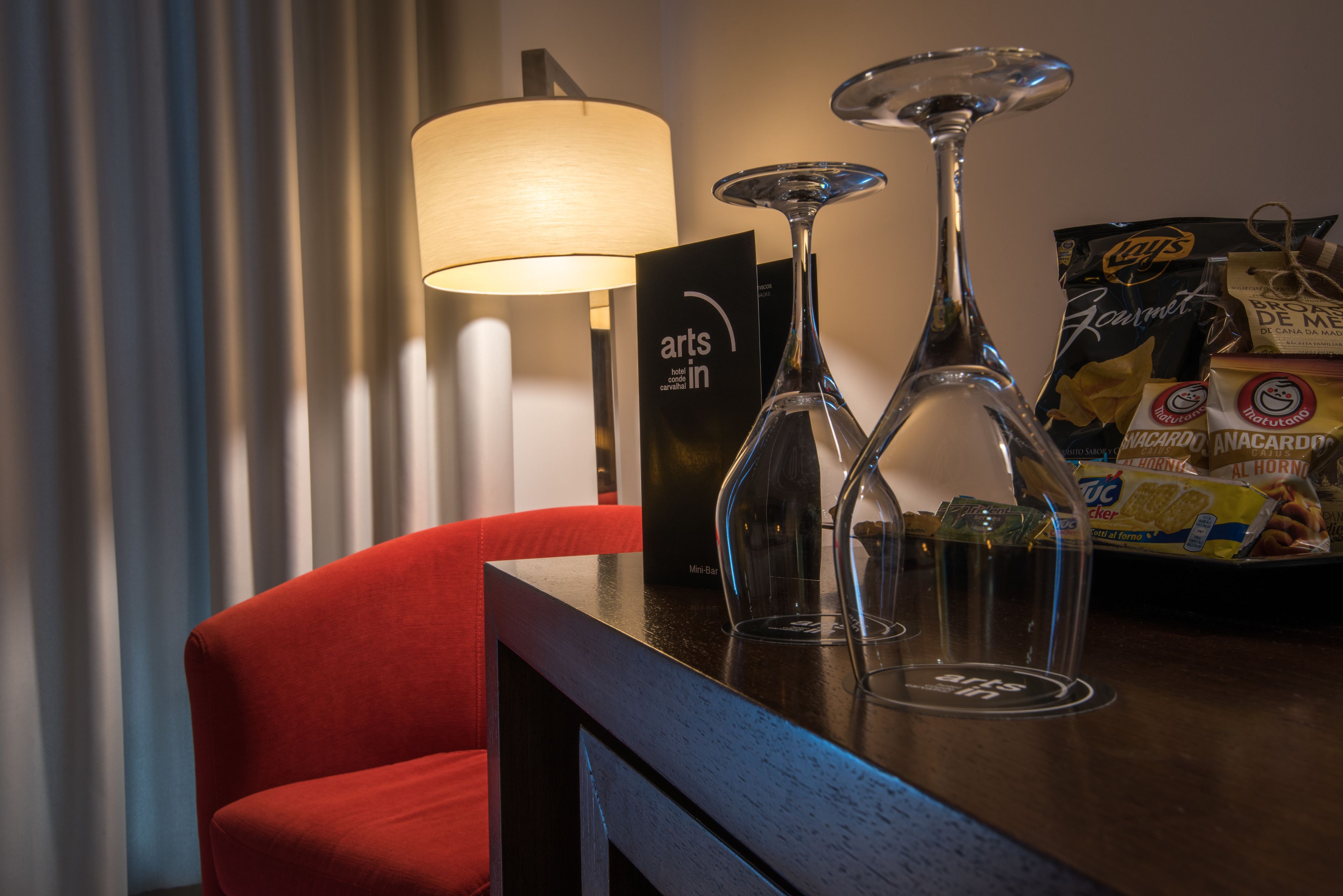 A hotel room with a red chair, a lamp, two wine glasses, and a minibar with snacks.