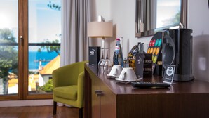 A hotel room with a wooden desk, a green armchair, a lamp, a minibar, a coffee machine, and a view of the sea.