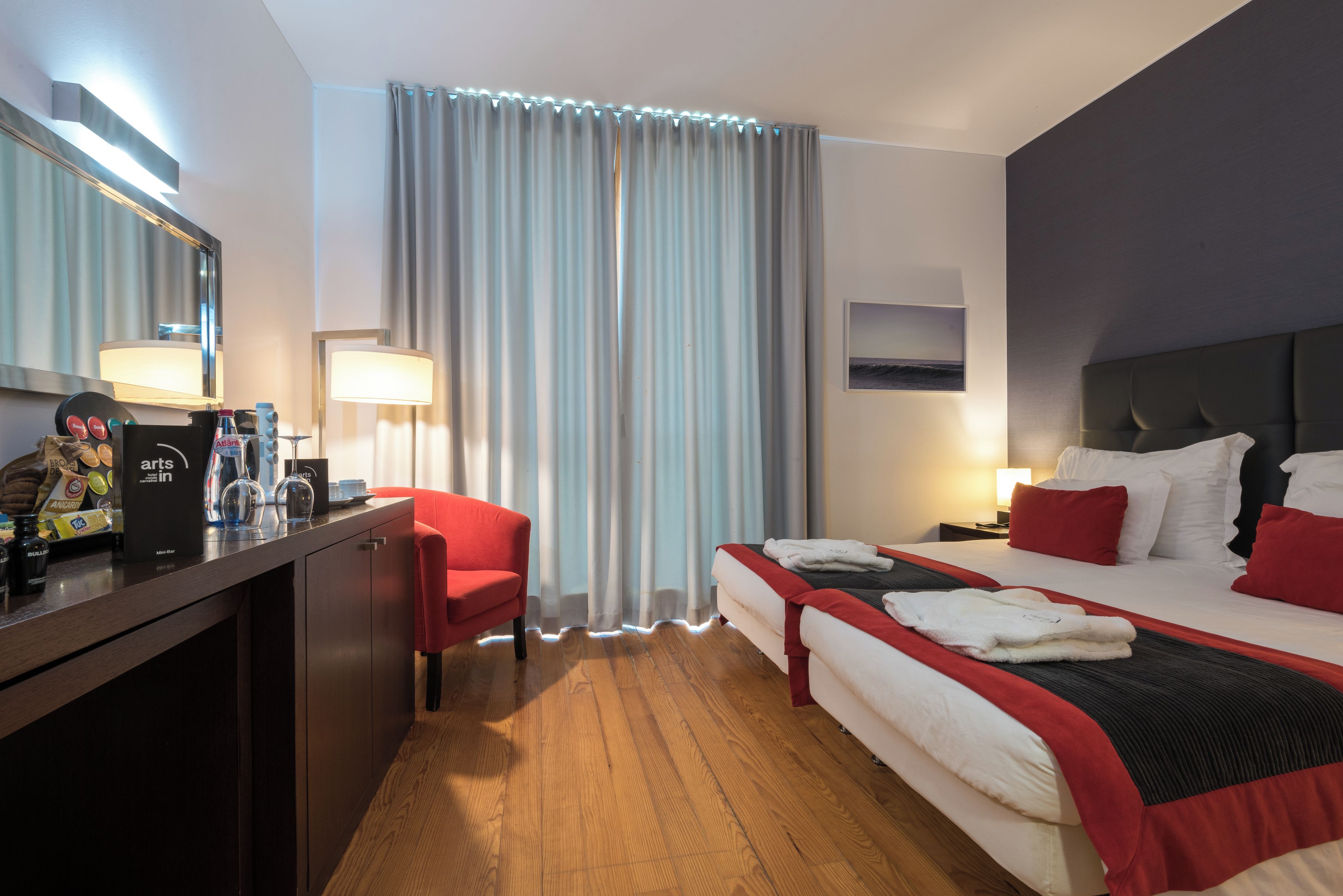 A hotel room with a desk, two beds, a minibar, and a large window with curtains.