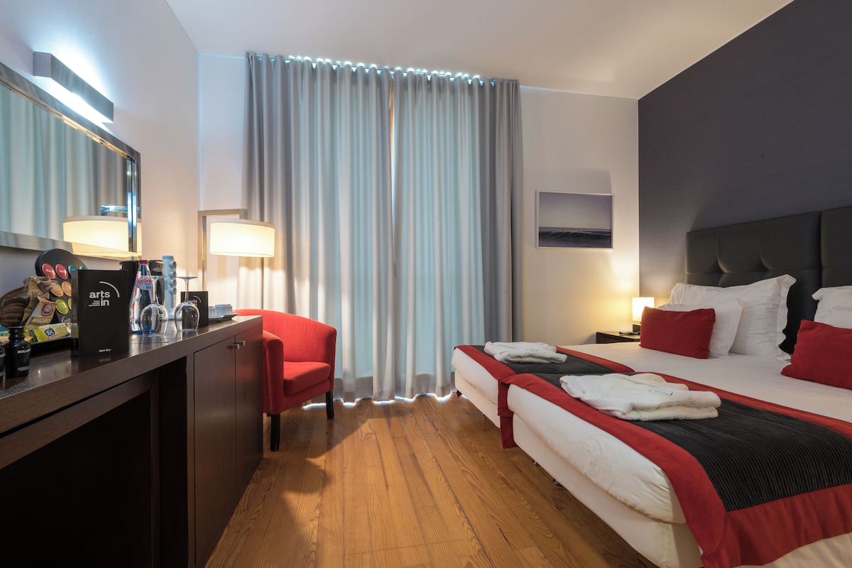 A hotel room with a wooden floor, a desk with a mirror, a red chair, a bed with white and red bedding, and a large window with curtains.