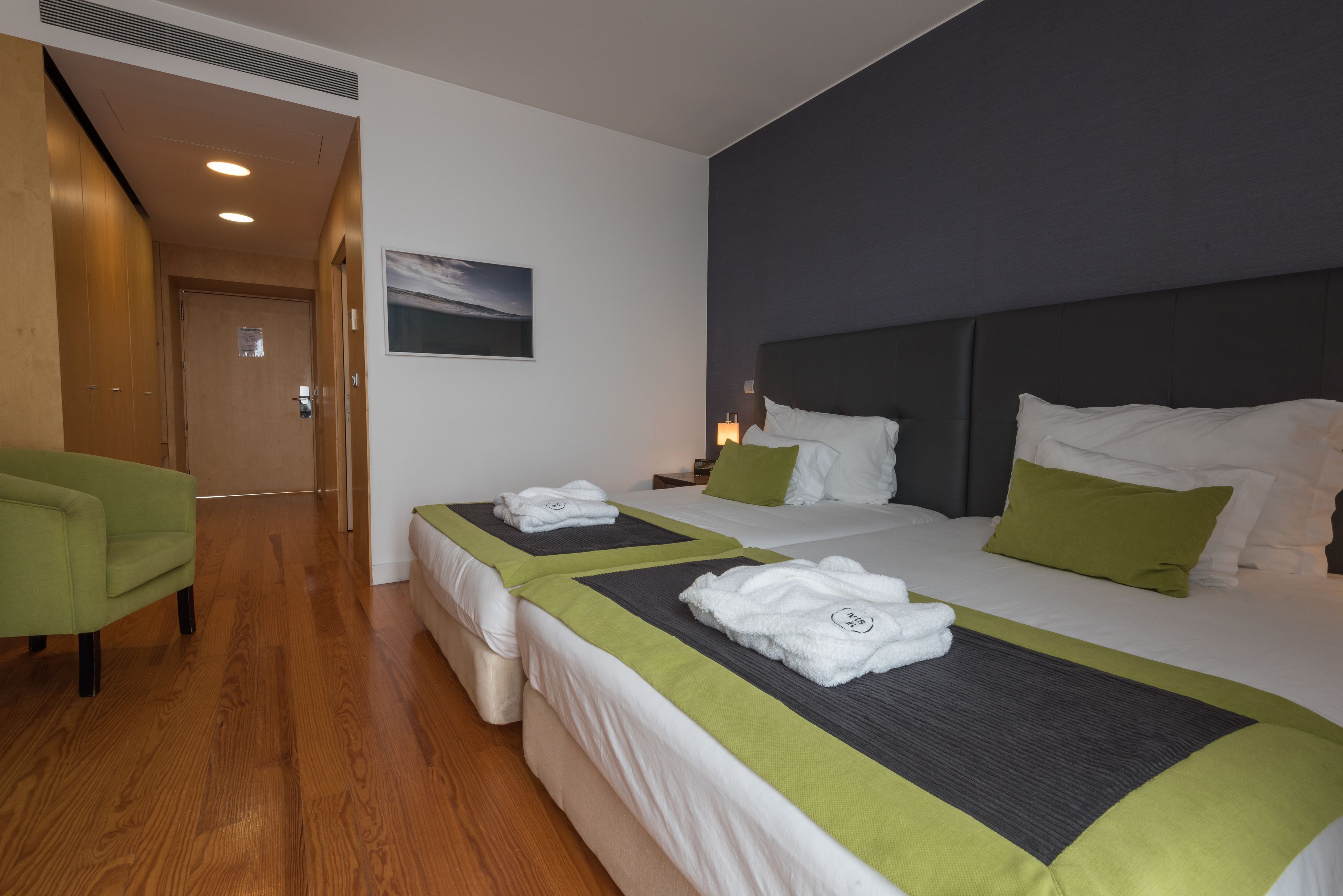 A hotel room with two beds, a green armchair, a wooden floor, and a painting on the wall.