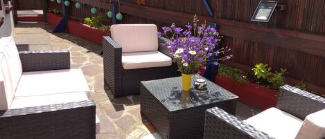 Un patio exterior con muebles de mimbre, una mesa pequeña con un florero y una valla de madera decorada.
