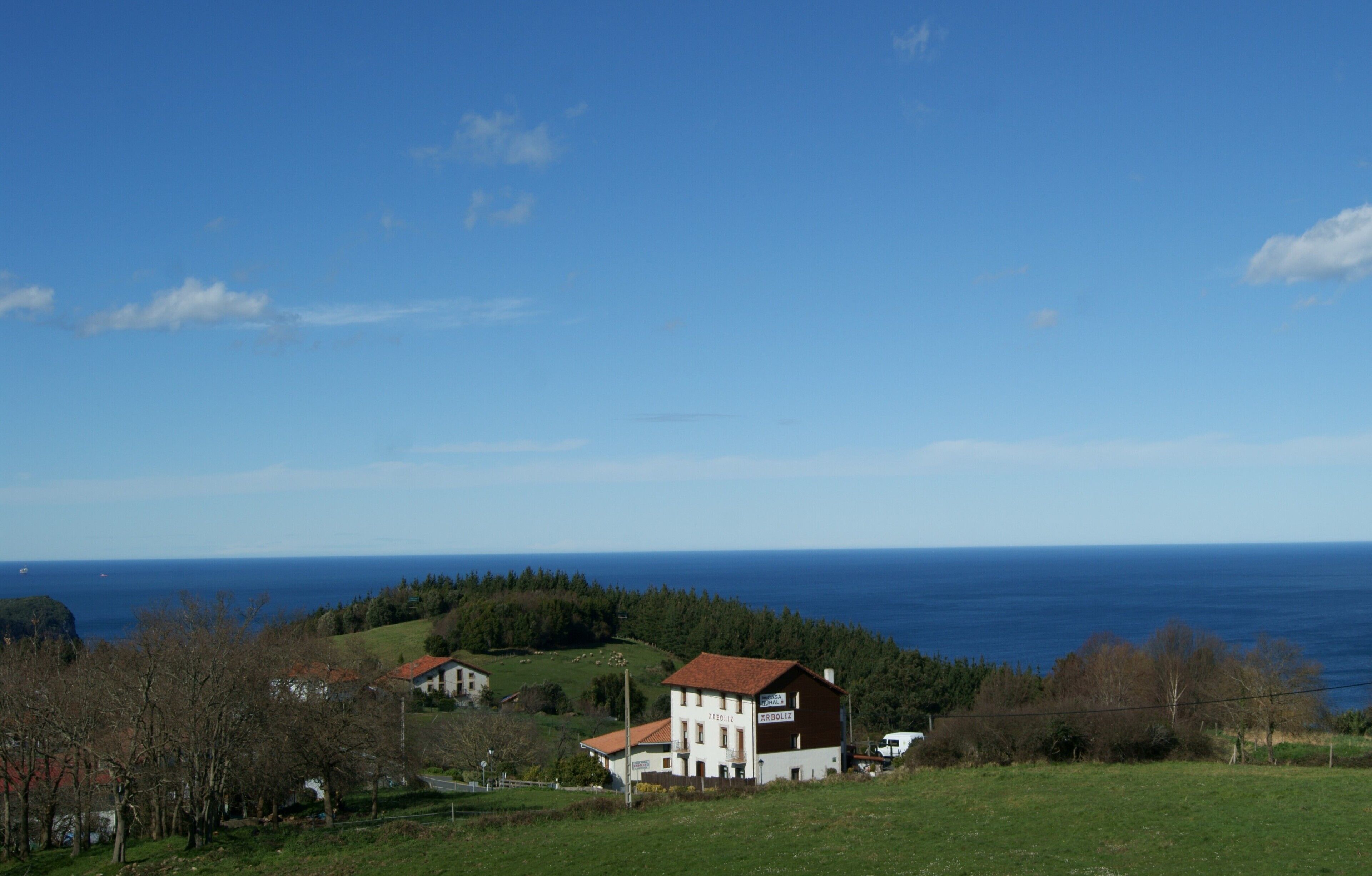 Et landhus med rødt tag, omgivet af grønne marker og udsigt til havet.