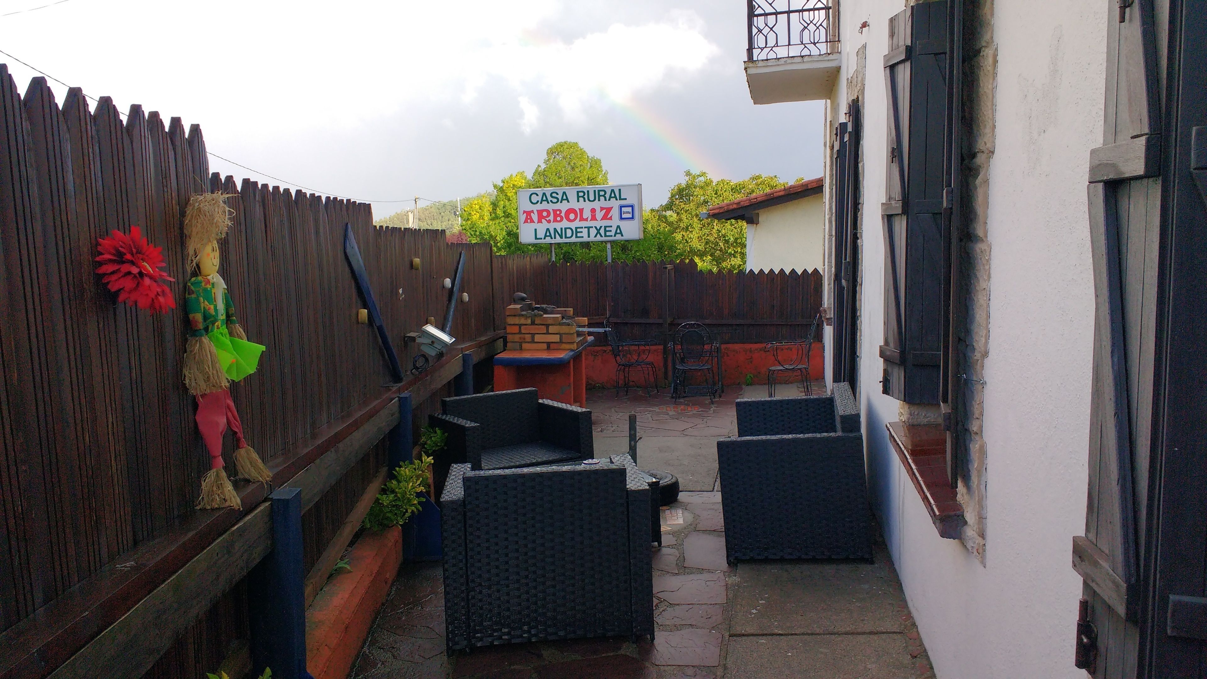 Un patio estrecho con valla de madera, mobiliario exterior y un cartel que dice 'Casa Rural Arbólez Landetxea'.