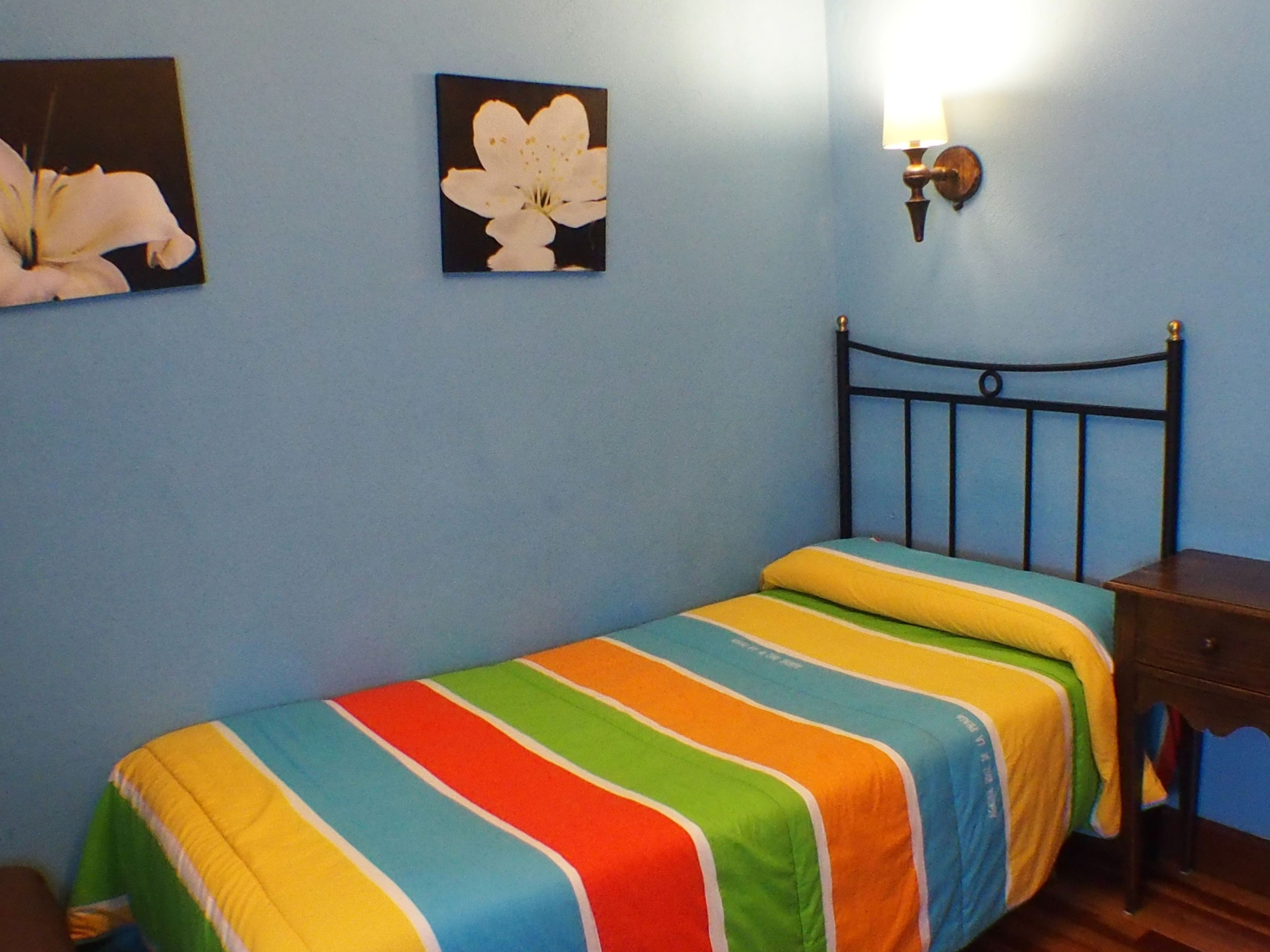 Un dormitorio con una cama individual, una mesita de noche, una lámpara de pared y dos cuadros enmarcados.