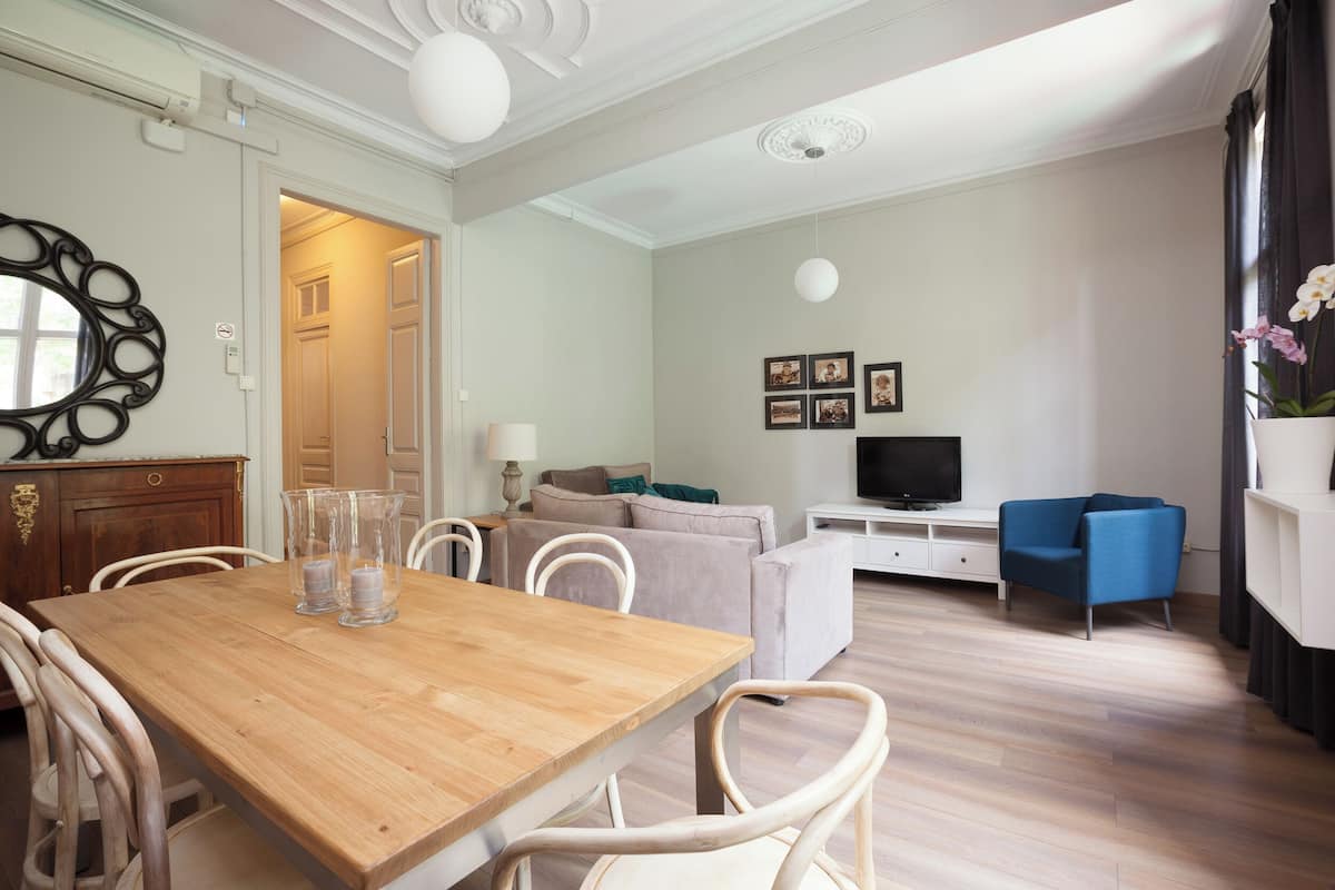 A living room with a wooden dining table, white chairs, a sofa, a TV, and a blue armchair.