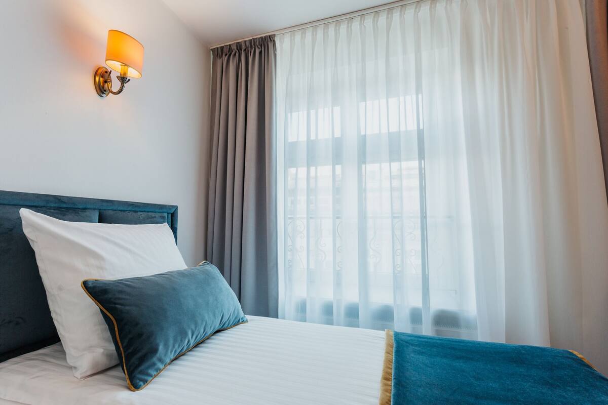 A hotel room with a bed, a wall-mounted lamp, and a large window with curtains.