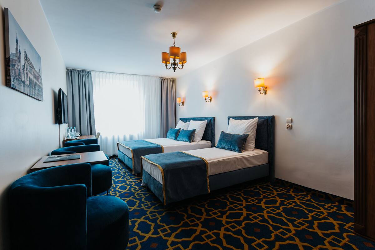 A hotel room with two beds, a desk, and a chair. There are two wall-mounted lamps and a chandelier.