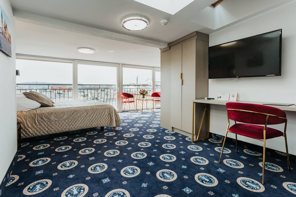 A hotel room with a bed, a desk, a TV, and a balcony with red chairs.