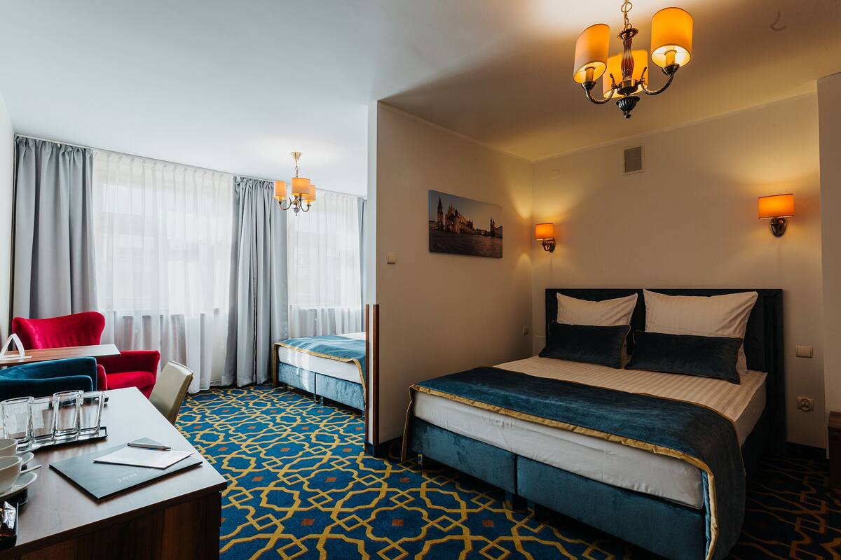 A hotel room with a large bed, a desk with a chair, a red armchair, and a window with sheer curtains.