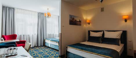 A hotel room with a large bed, a desk with a chair, a red armchair, and a window with sheer curtains.