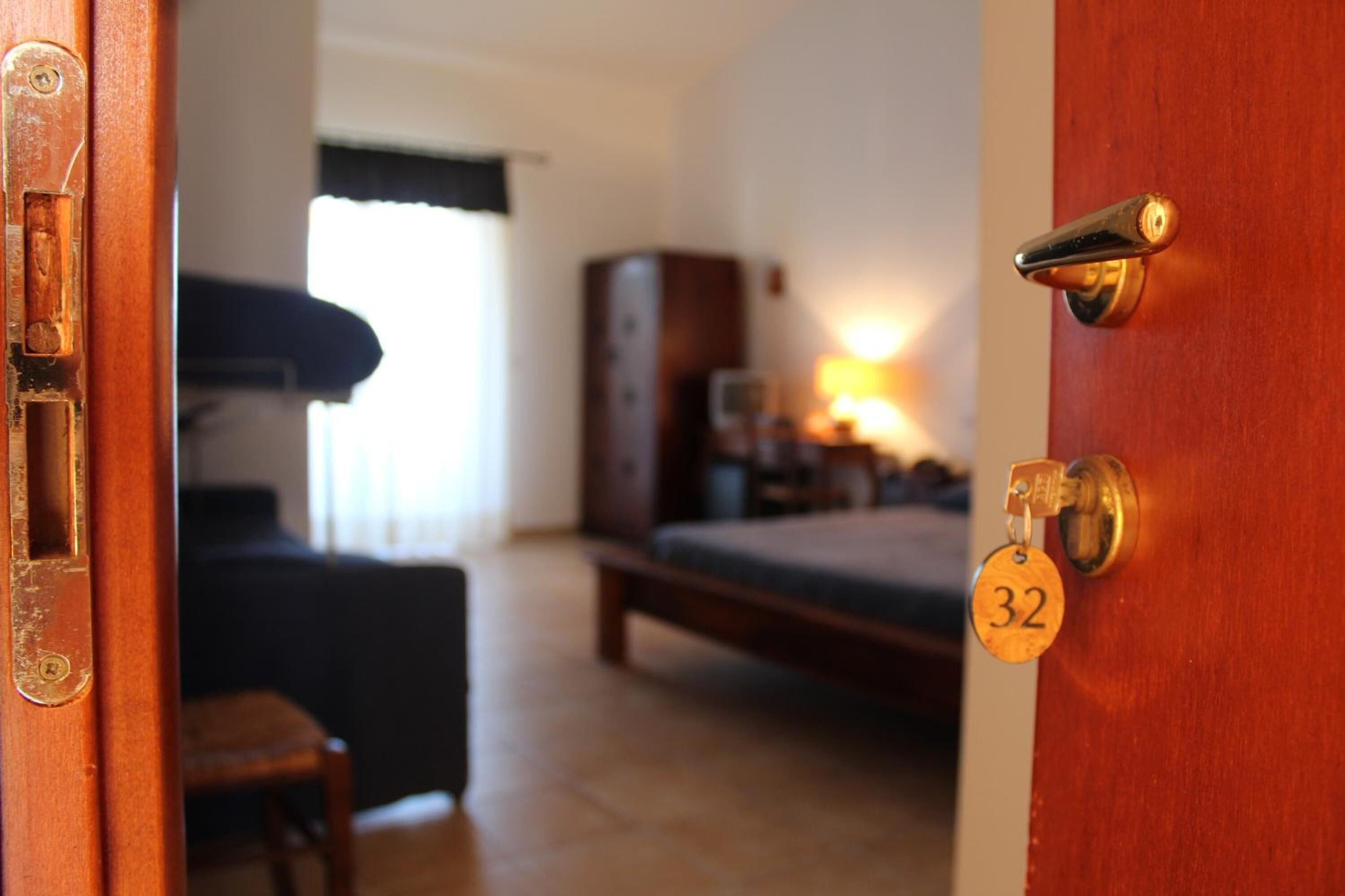 A wooden door with a brass handle and a keyhole, a number 32 key tag, a bed, a nightstand, a lamp, and a window with curtains.
