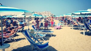Une plage bordée de rangées de transats et de parasols, un ciel dégagé et une vue lointaine sur des bâtiments.
