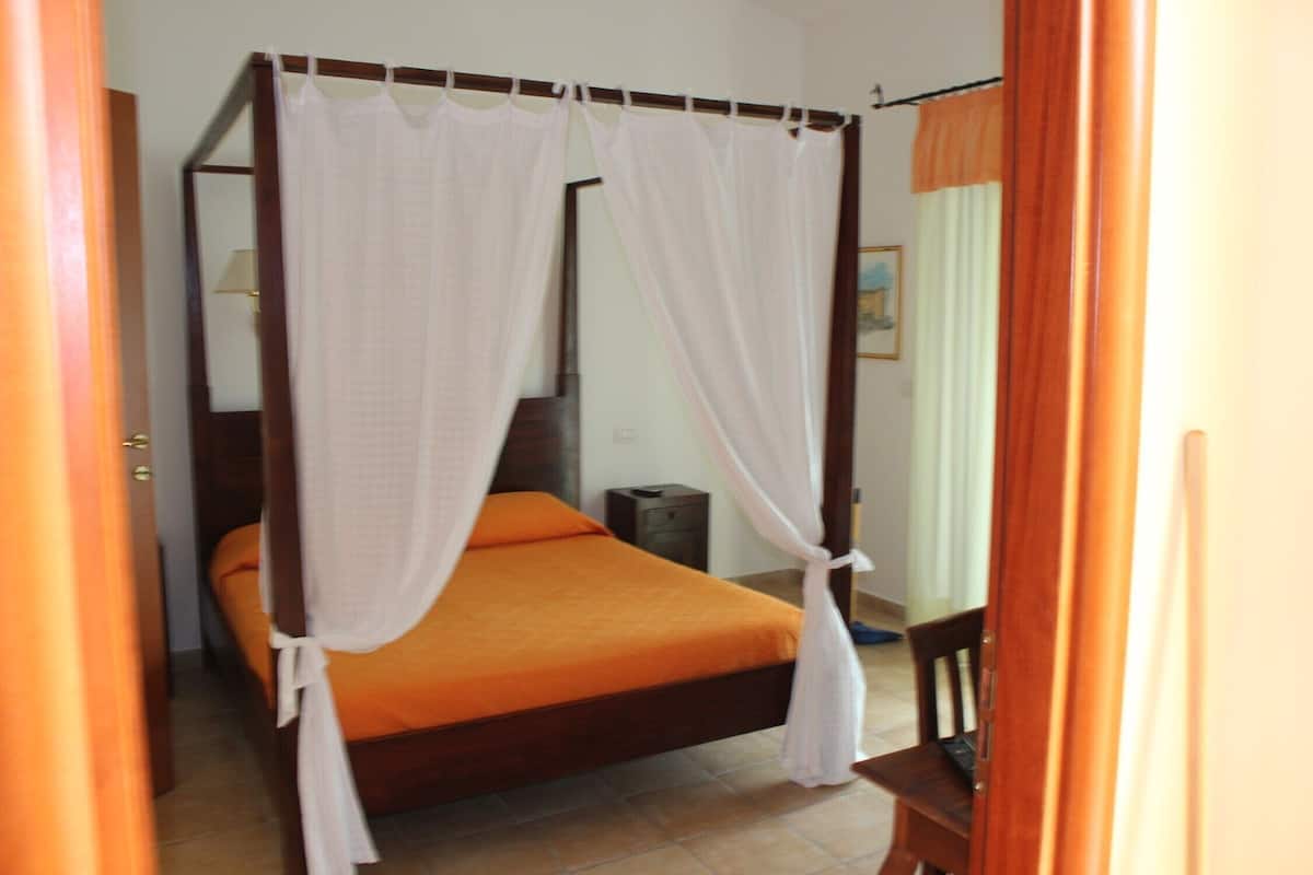 Ein Bett mit Himmel, weiße Vorhänge, eine orangefarbene Bettdecke und ein hölzernes Kopfteil.