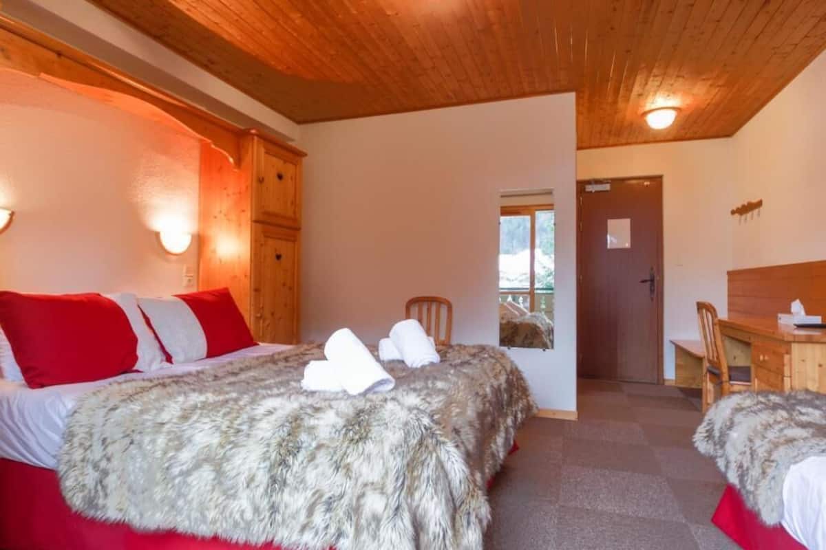 Ein Zimmer mit zwei Betten, jeweils mit roter und weißer Bettwäsche, einem Fellüberwurf und einem Holz-Kopfteil. Es gibt einen Holztisch mit Stuhl und eine Tür, die in einen anderen Raum führt.