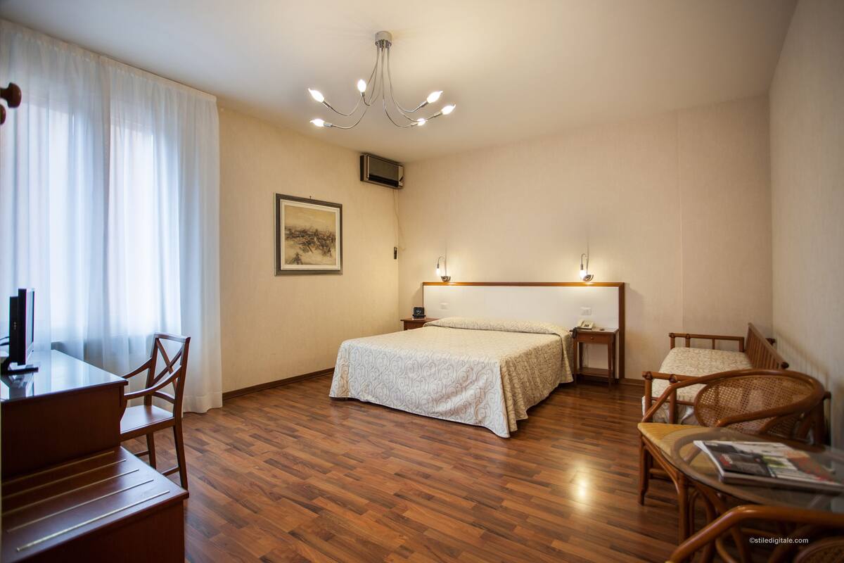 Una habitación de hotel con piso de madera, una cama con cabecera, un escritorio con silla, una mesita con una silla y una araña.