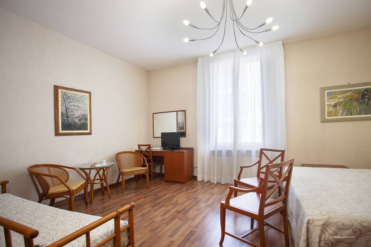 Habitación de hotel con piso de madera, un escritorio con computadora, dos sillas, una cama y una araña.