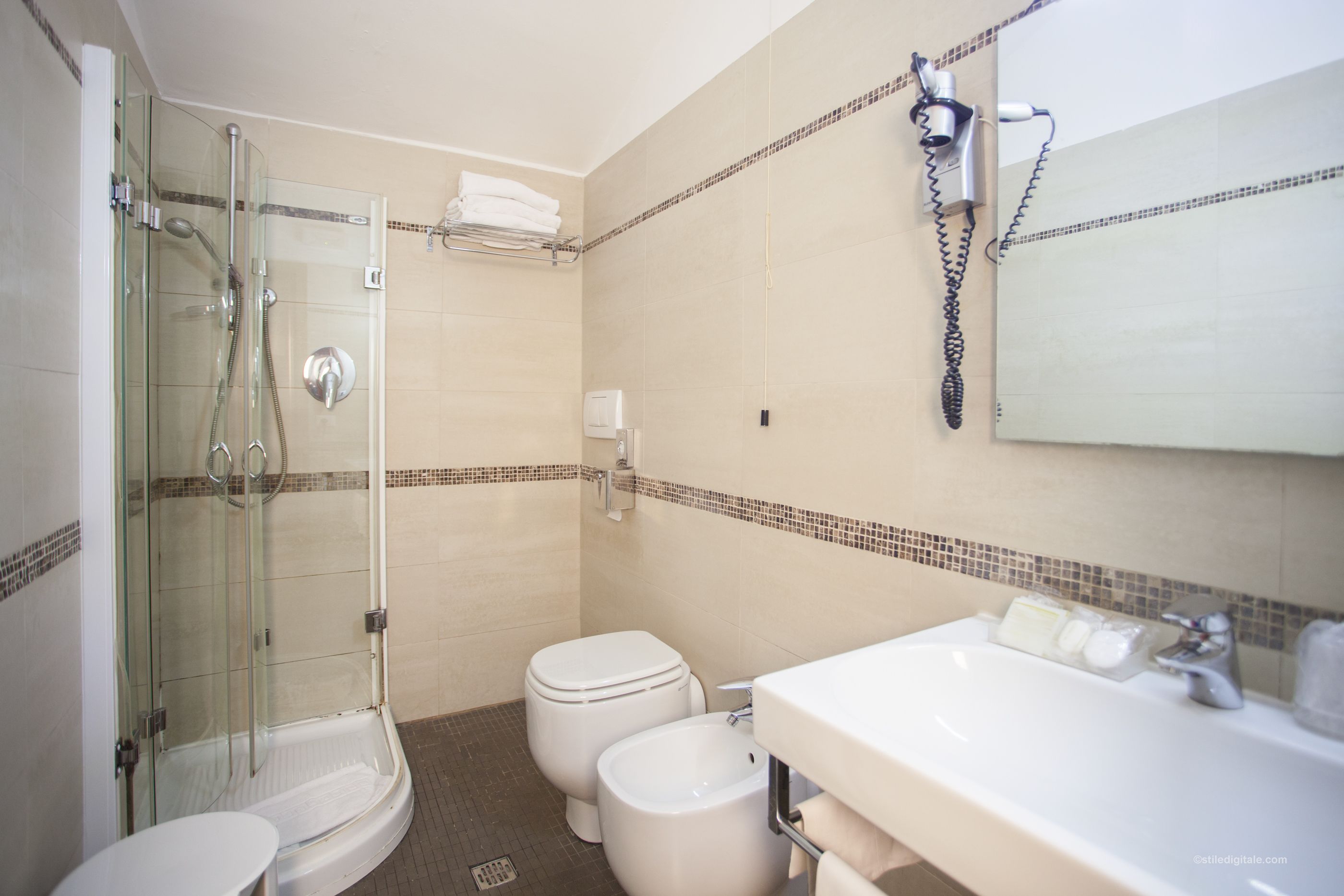 Une salle de bain avec une douche à parois de verre, deux toilettes, un lavabo et un sèche-cheveux fixé au mur.