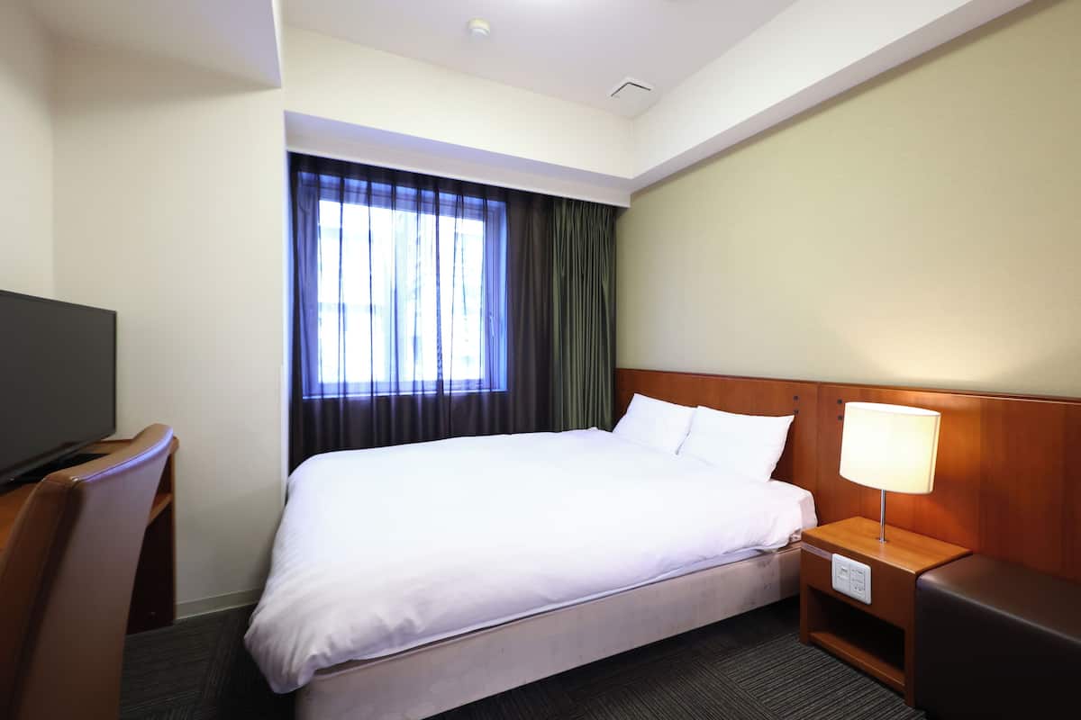A hotel room with a bed, a TV, a nightstand with a lamp, and a window with curtains.