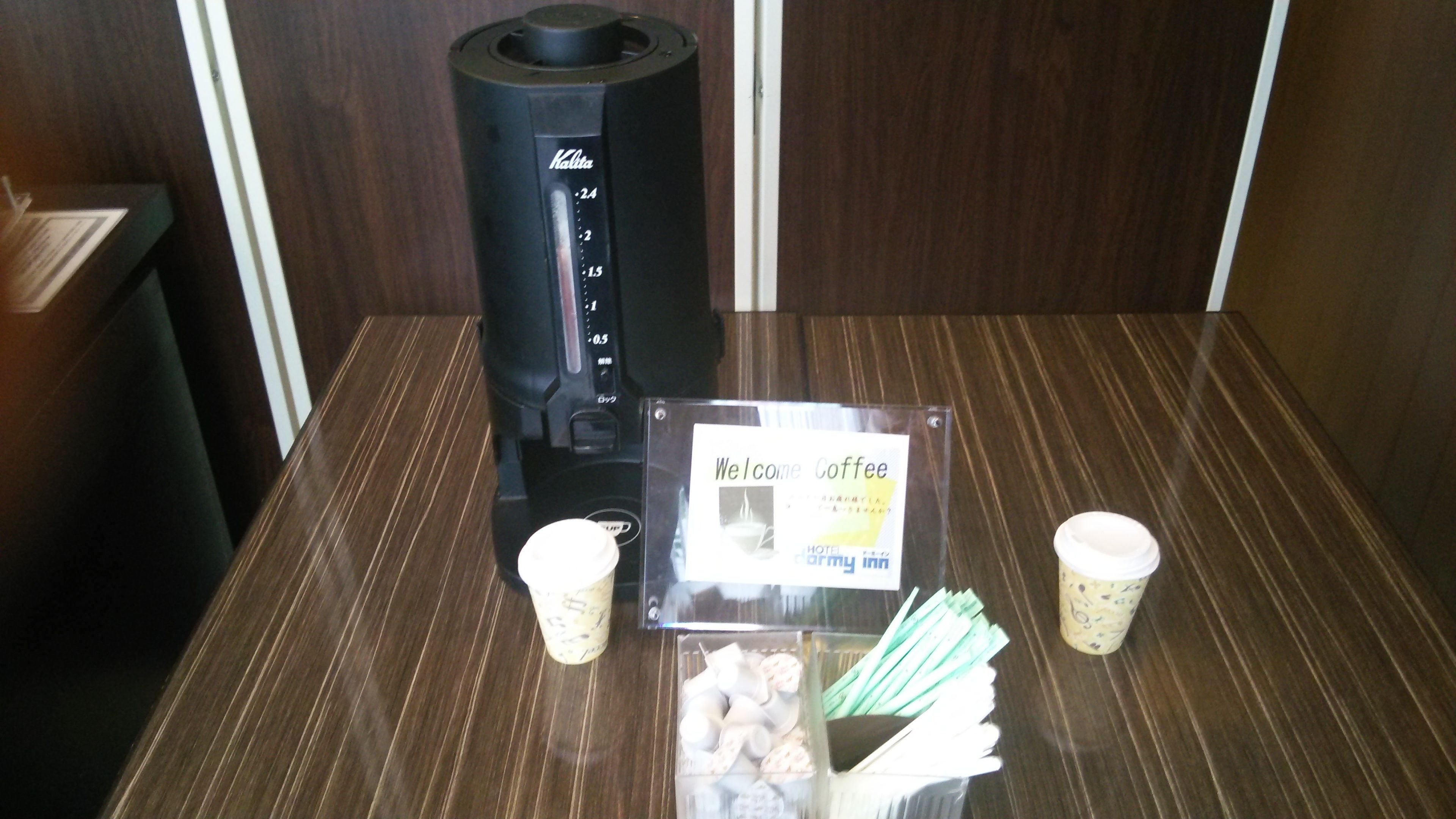 A coffee machine, a sign reading "Welcome Coffee", and two disposable cups on a table.