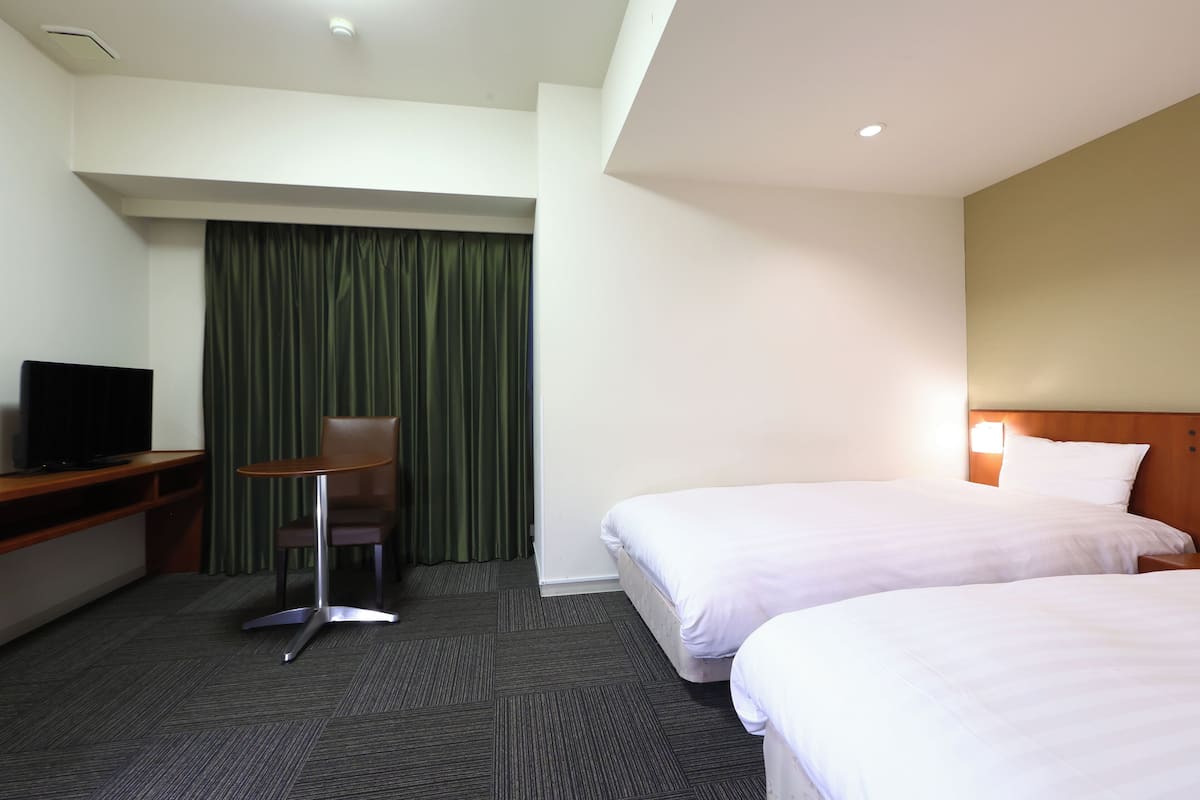 A hotel room with two beds, a desk with a chair, a television, and a green curtain.