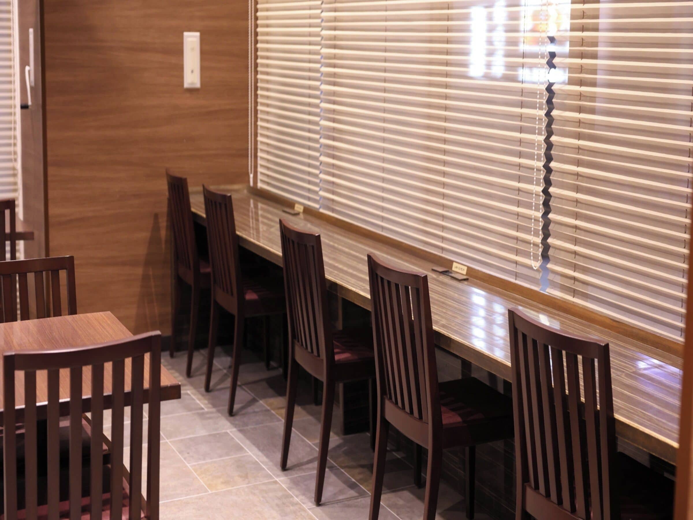 A dining area with wooden tables and chairs, a tiled floor, and a window with blinds.