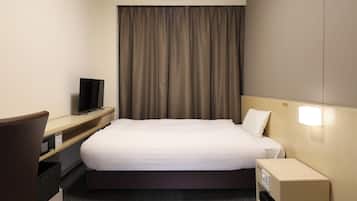 A hotel room with a bed, a desk with a television, a nightstand, and a wall-mounted light.