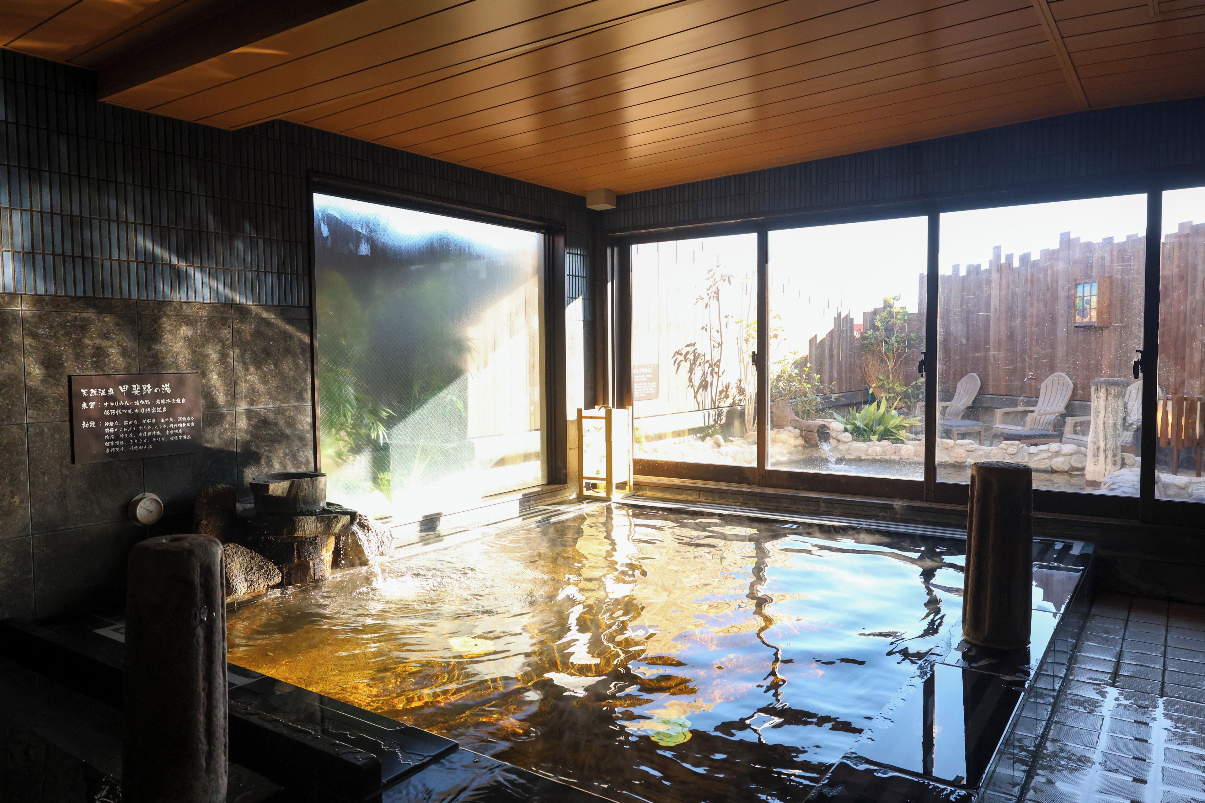 A hot spring bath area with a wooden ceiling, stone walls, and a view of a wooden fence outside.