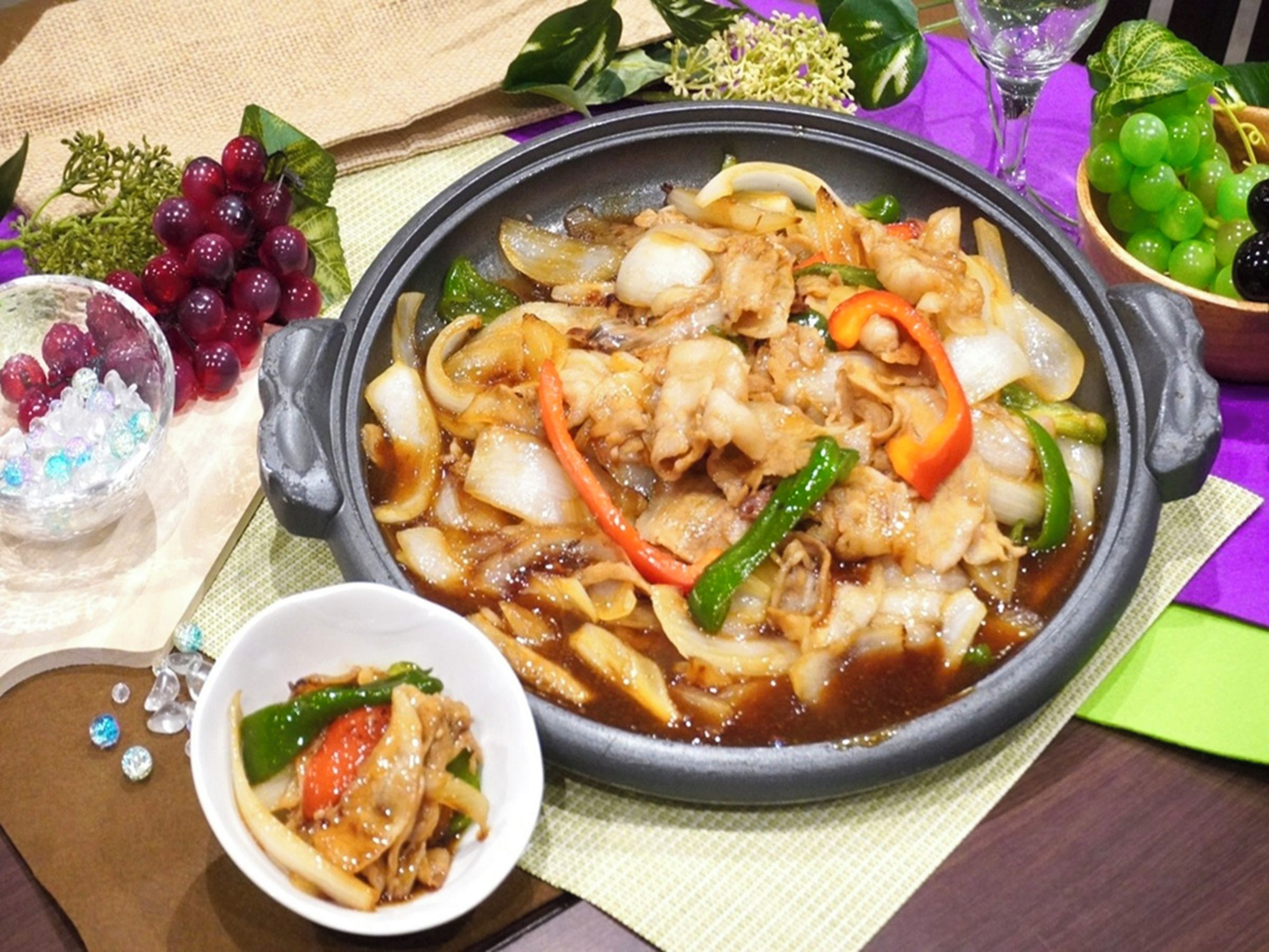 A meal with various vegetables and meat in a black pan, accompanied by a bowl of sauce and a glass of water.