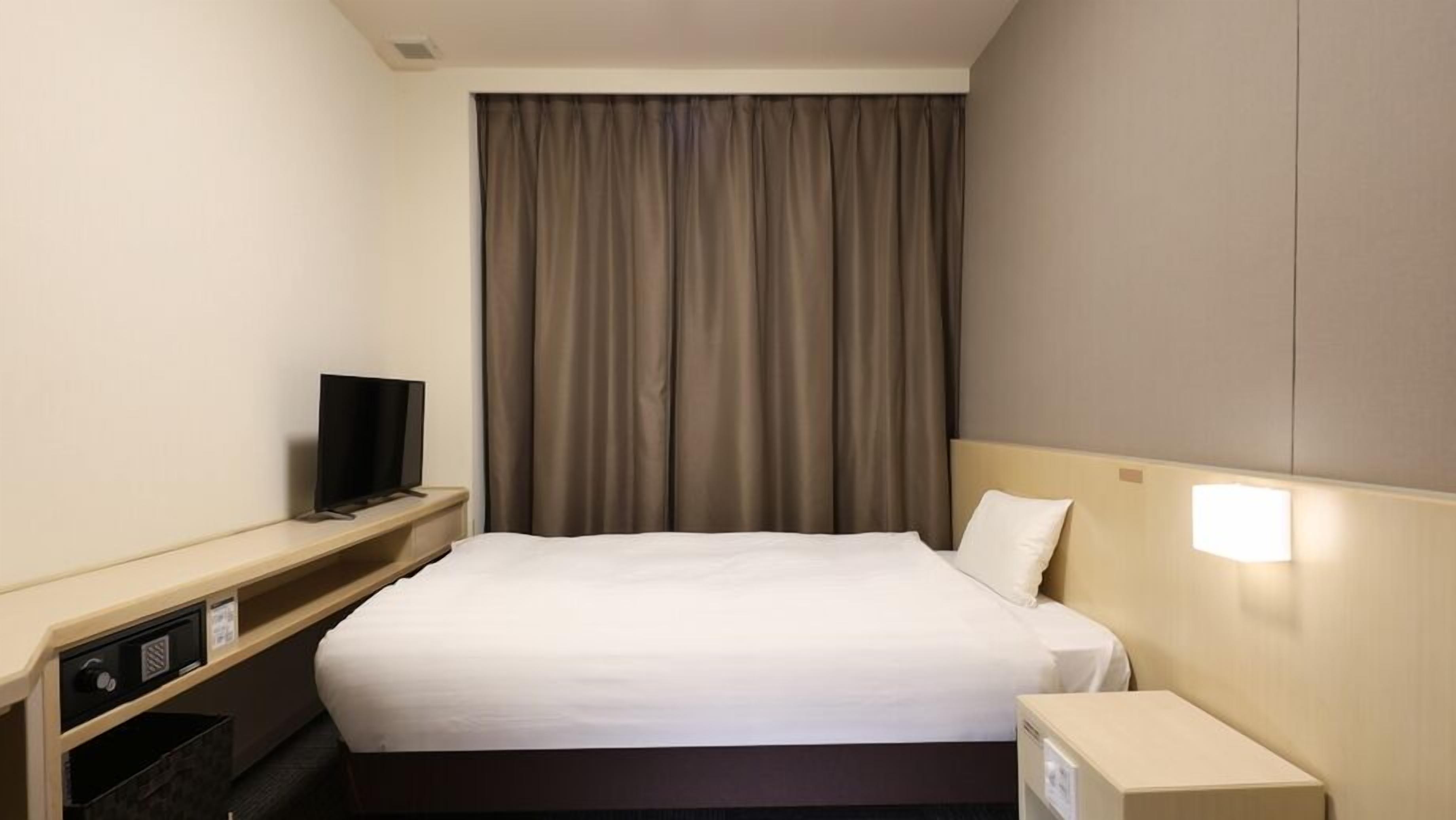 A hotel room with a bed, a television on a stand, a nightstand with a lamp, and a window with curtains.
