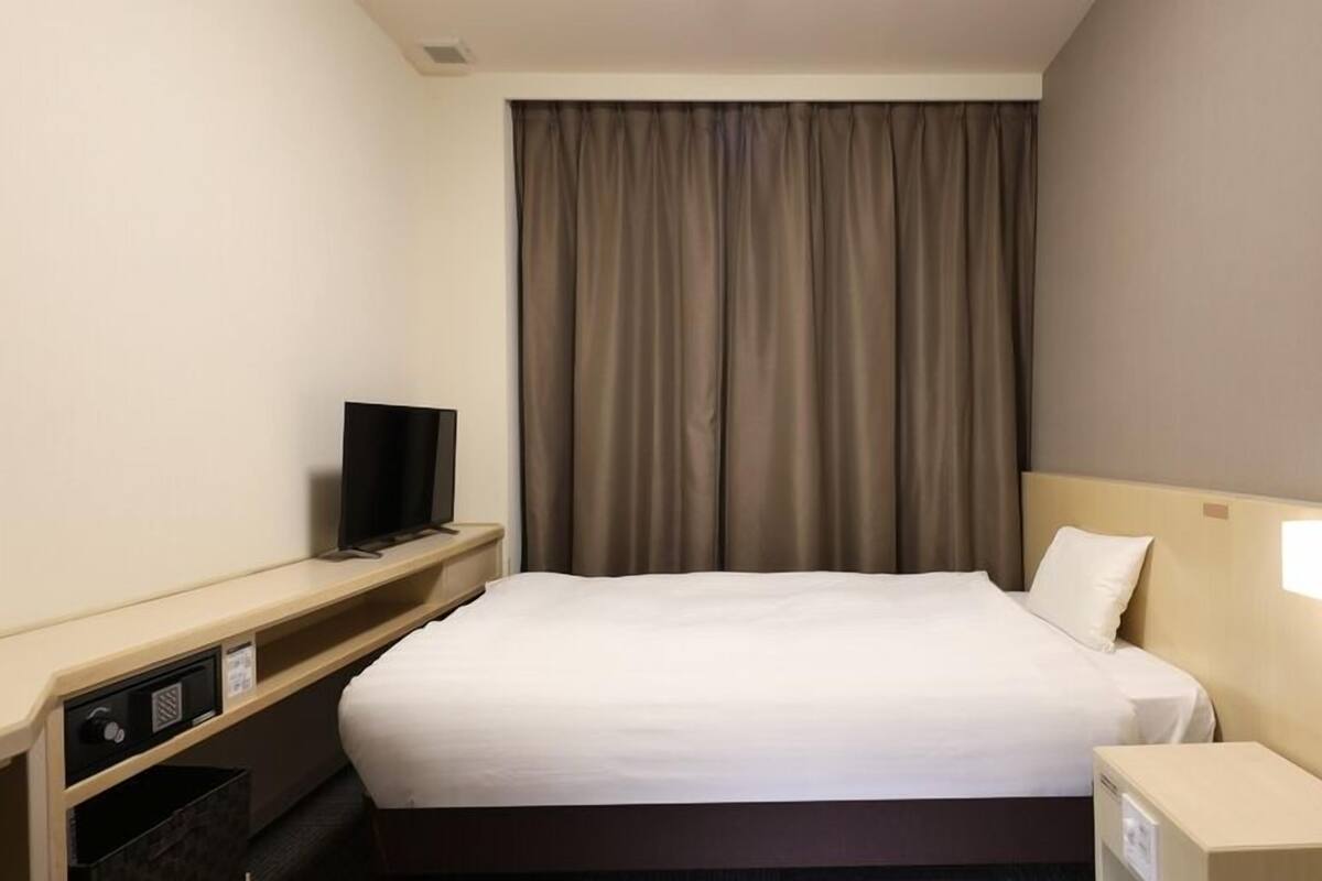 A hotel room with a bed, a television on a stand, a nightstand with a lamp, and a window with curtains.