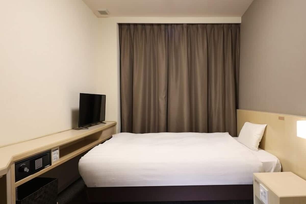 A hotel room with a bed, a television on a stand, a nightstand with a lamp, and a window with curtains.