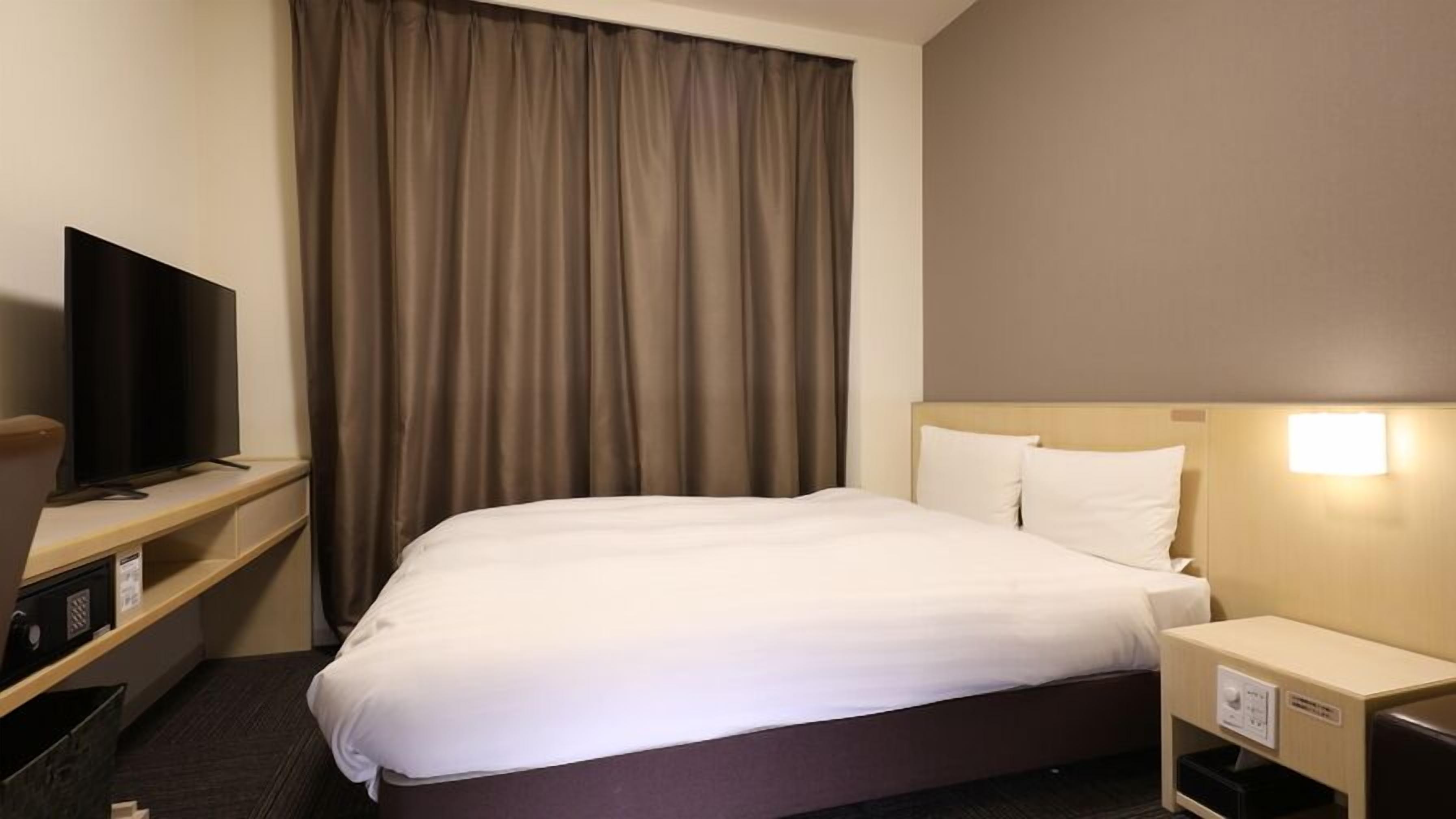 A hotel room with a bed, a television on a stand, a nightstand, and a window with curtains.