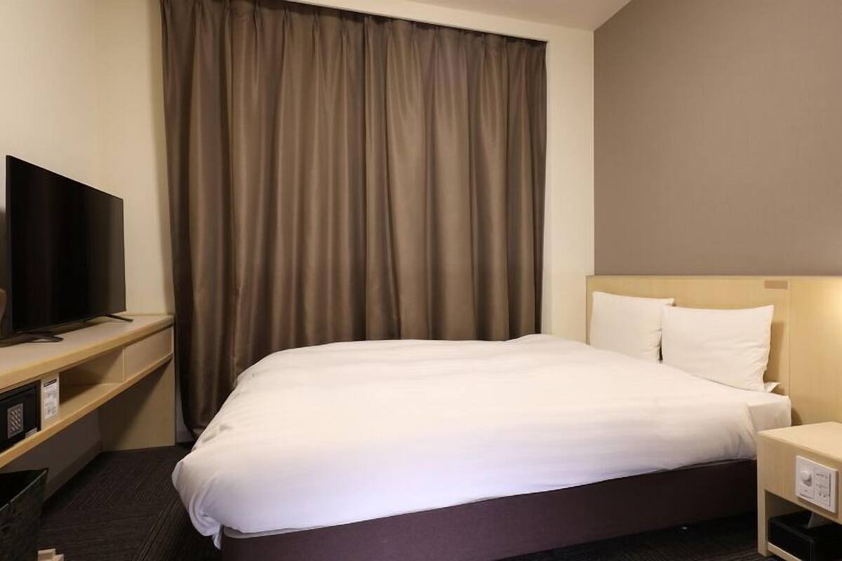 A hotel room with a bed, a television on a stand, a nightstand, and a window with curtains.