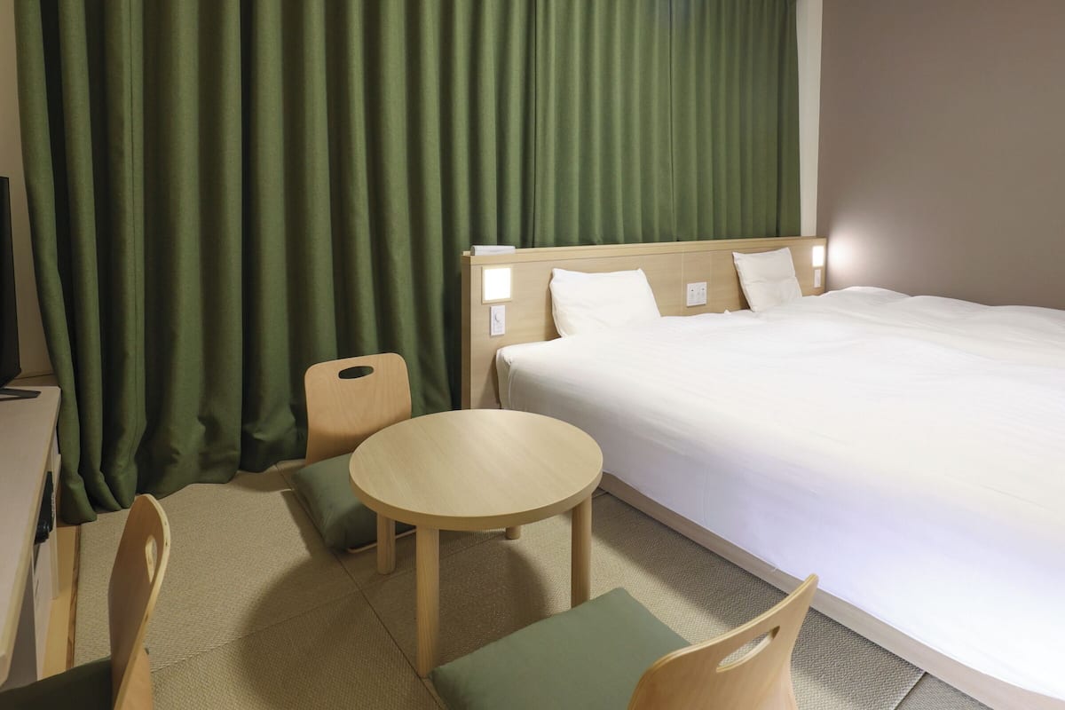 A hotel room with a bed, a small round table, two chairs, a desk, and a green curtain.