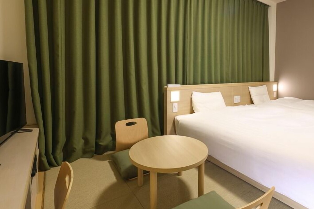 A hotel room with a bed, a small round table, a chair, a desk with a computer, and green curtains.