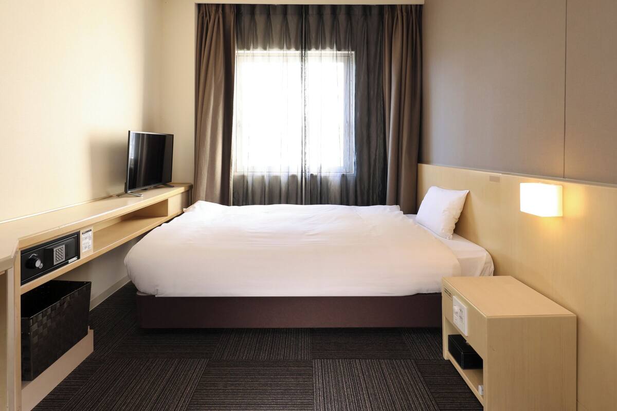 A hotel room with a large bed, a TV on a wooden stand, a bedside table, and a window with curtains.