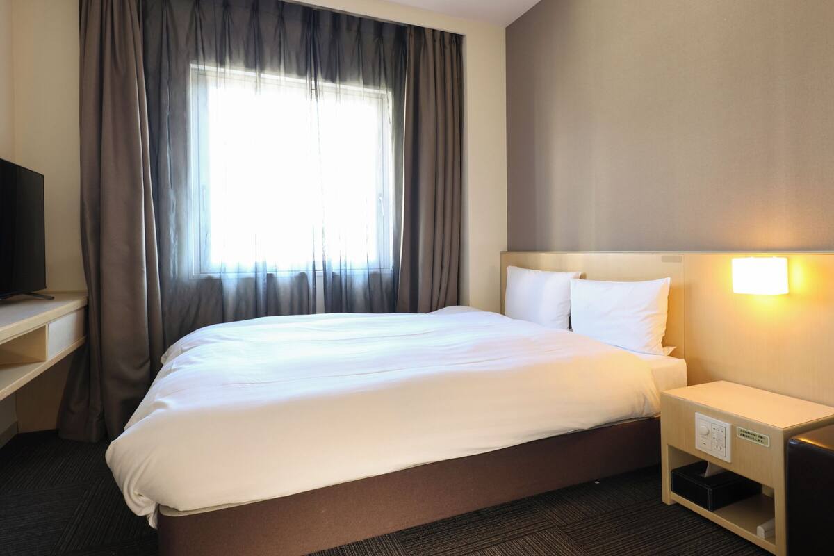 A hotel room with a large bed, a bedside table with a lamp, a TV on a stand, and a window with curtains.