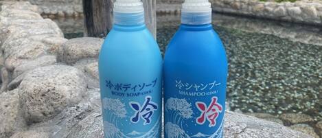 Dos botellas de jabón corporal y champú con texto en japonés, sobre una superficie de piedra cerca de un estanque.