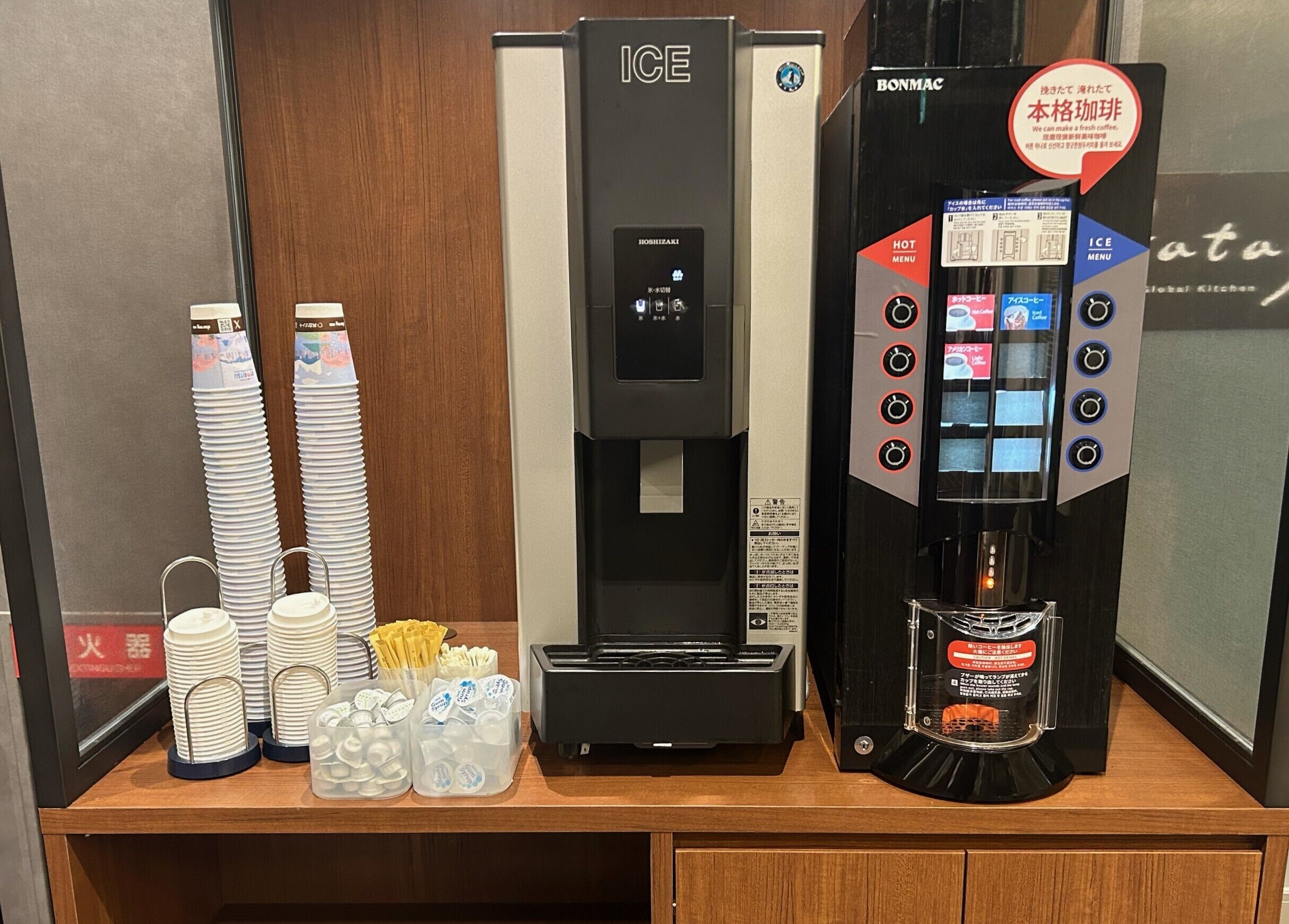 A coffee machine with a digital display and a menu, a stack of disposable cups, and a container with coffee pods.