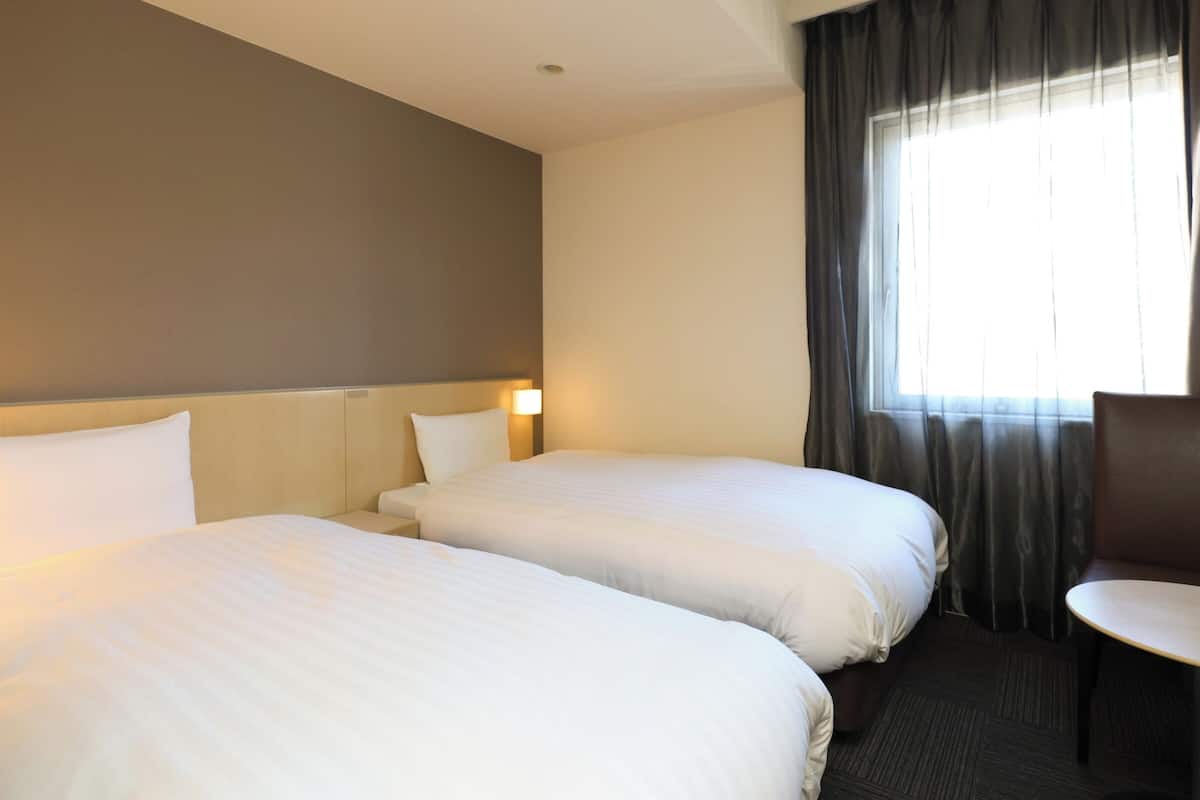 A hotel room with two beds, a small table, a chair, and a window with curtains.
