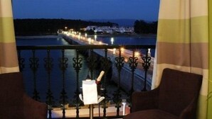 Un balcón con vista a un muelle iluminado que se extiende sobre el agua, una silla y una pequeña mesa con una lámpara.