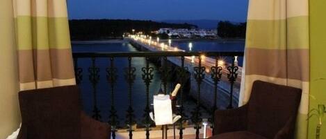 A balcony with a view of a lit pier extending into a body of water, a chair, and a small table with a lamp.