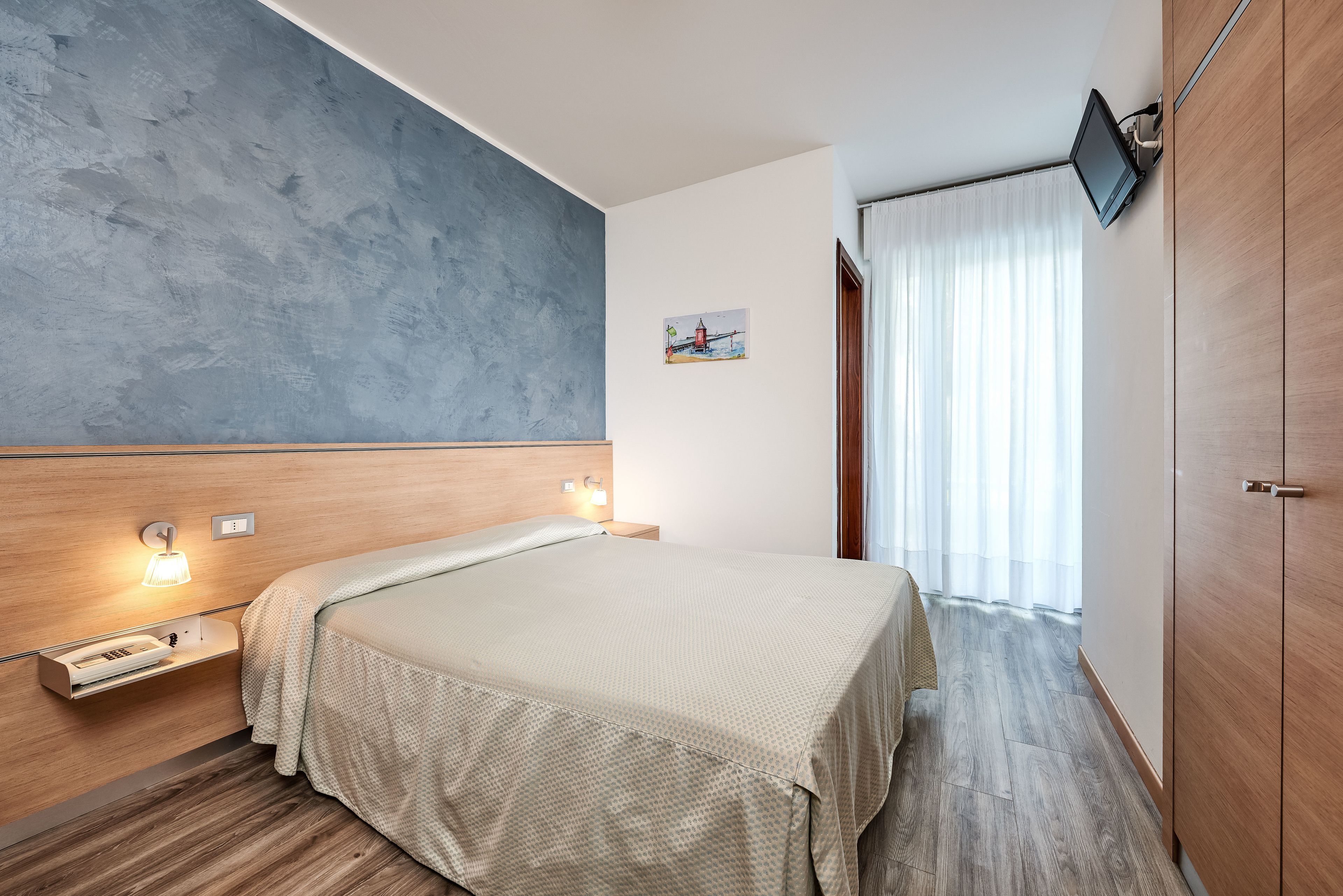 Habitación de hotel con cama, mesita de noche con lámpara, una obra de arte pequeña en la pared y un televisor montado.