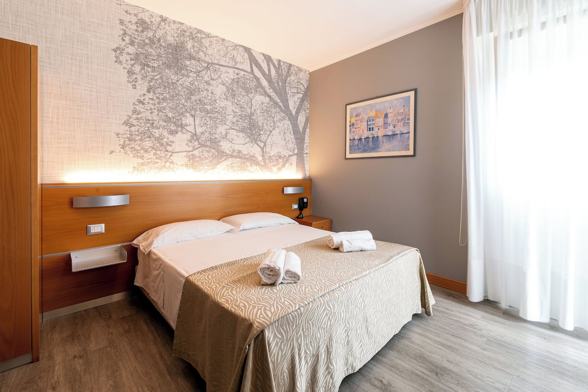 A hotel room with a large bed, a headboard with a tree mural, a bedside table, a nightstand, a wall-mounted lamp, a painting, and a window with curtains.
