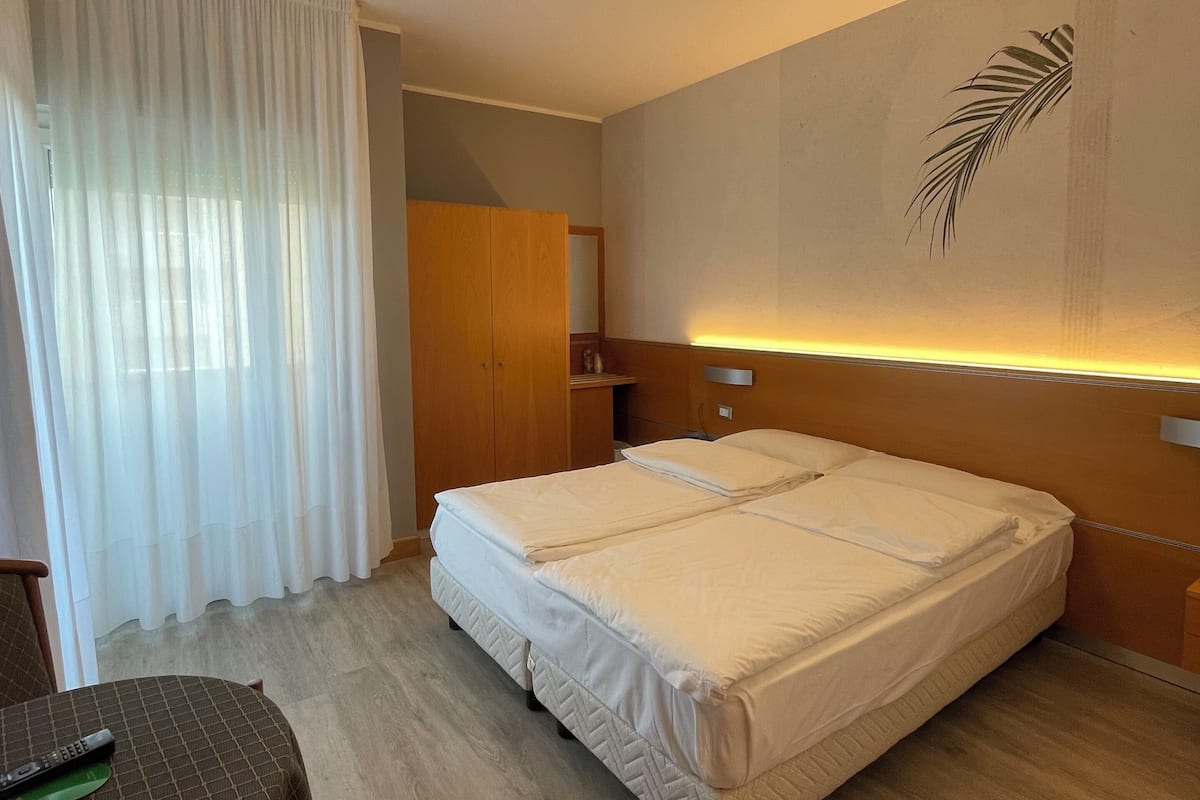 A hotel room with a bed, a small table, a wooden wardrobe, and a wall-mounted light fixture.