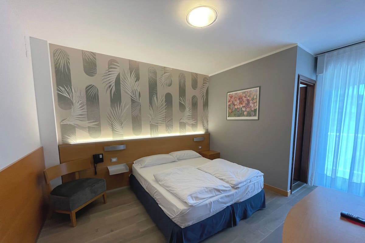 A hotel room with a bed, a desk, a chair, a nightstand, a wall with a leaf pattern, a painting, and a window with curtains.